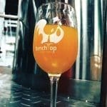 A tall stemmed glass of orange beer etched with the BenchTop Brewing Company logo, set on a steel brewery floor.