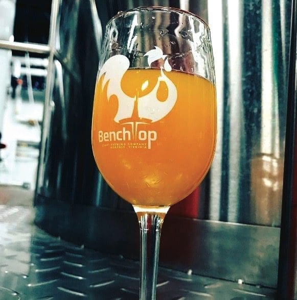 A tall stemmed glass of orange beer etched with the BenchTop Brewing Company logo, set on a steel brewery floor.