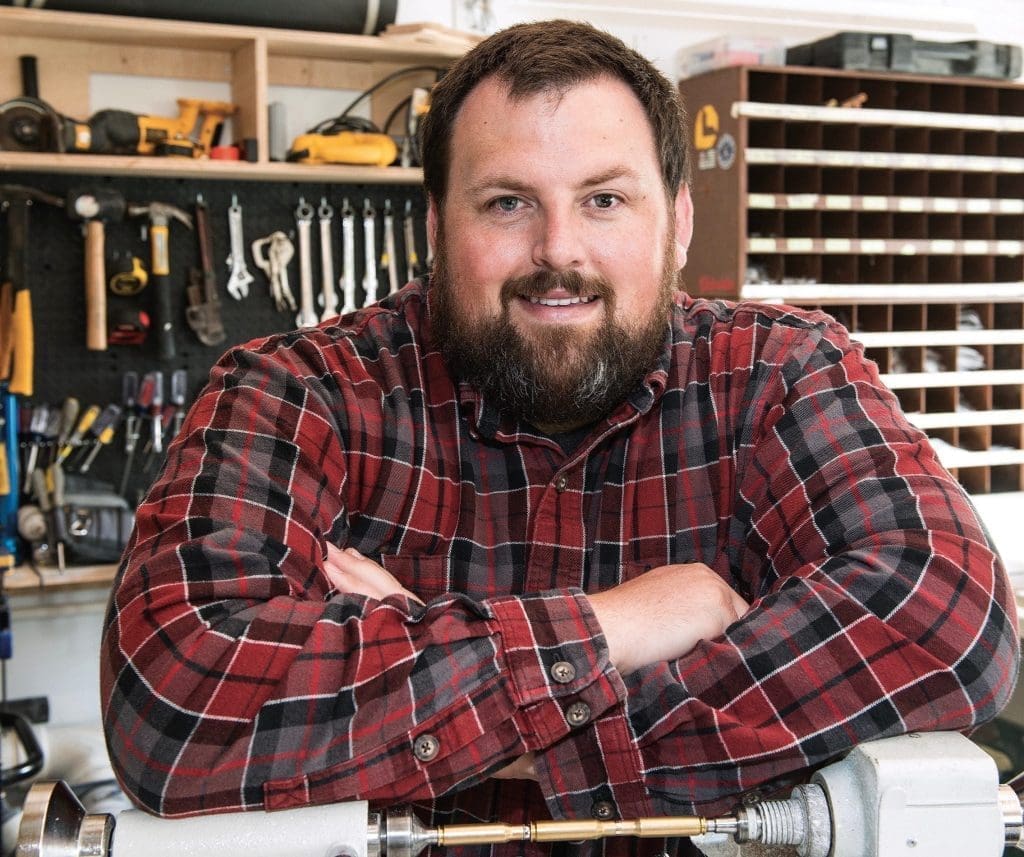 A bearded man in a red plaid flannel shirt leans with folded arms on a workbench in a workshop lined with tools.