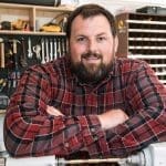 A bearded man in a red plaid flannel shirt leans with folded arms on a workbench in a workshop lined with tools.