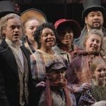 A cast of costumed performers in Victorian dress, including top hats and plaid shawls, gathers closely on a dark stage.