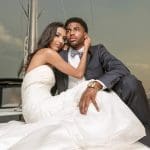 A bride in a white strapless gown and a groom in a gray suit with a checked bow tie pose on a sailboat deck.