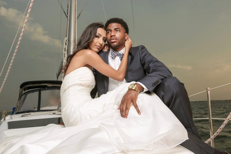 A bride in a white strapless gown and a groom in a gray suit with a checked bow tie pose on a sailboat deck.