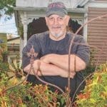 A bearded man in a dark cap and apron stands arms crossed behind a wrought-iron trellis of red and yellow peppers.