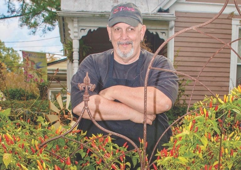 A bearded man in a dark cap and apron stands arms crossed behind a wrought-iron trellis of red and yellow peppers.