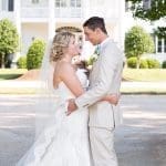 A bride in a lace gown and veil embraces a groom in a tan suit before a large white colonial mansion.