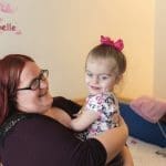 A woman with red hair and glasses smiles while holding a young girl with a pink bow in a bedroom with a name sign reading "Annabelle.