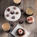 Overhead view of chocolate truffles on a white stand, beers, a red velvet cupcake, and a chocolate-glazed donut with cherry.