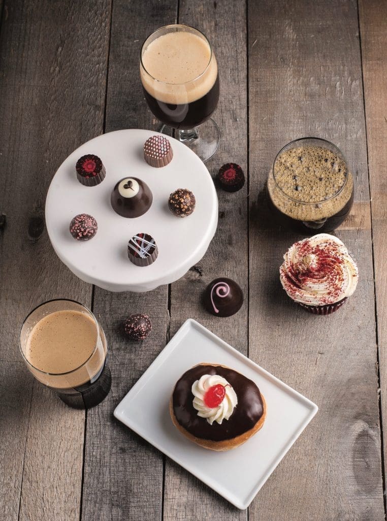 Overhead view of chocolate truffles on a white stand, beers, a red velvet cupcake, and a chocolate-glazed donut with cherry.