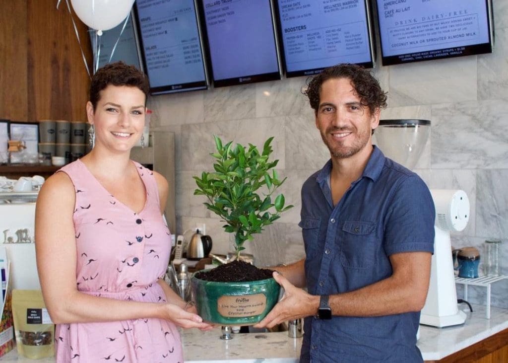 Two smiling people in a café hold a green pot with a small leafy citrus tree and a "Fruitive" plaque.