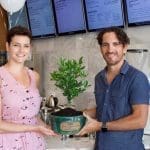 Two smiling people in a café hold a green pot with a small leafy citrus tree and a "Fruitive" plaque.