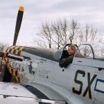 A man sits in the cockpit of a vintage silver fighter plane with a yellow-checkered nose and white star insignia.