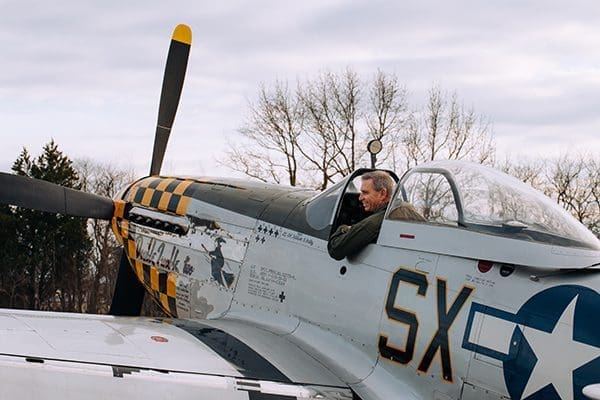 A man sits in the cockpit of a vintage silver fighter plane with a yellow-checkered nose and white star insignia.