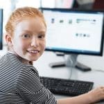 Young girl with red hair using computer at elementary school. Happy female child learning to use internet in a computer room.
