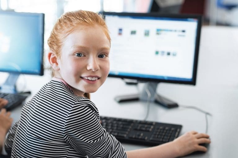 Young girl with red hair using computer at elementary school. Happy female child learning to use internet in a computer room.
