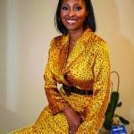Smiling woman in a yellow leopard-print suit with a black belt, seated on an orange chair.