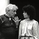 Black-and-white photo of an older man with white hair gesturing as he speaks to a woman with dark curly hair in a blouse.