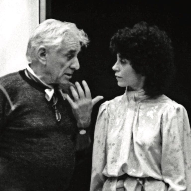 Black-and-white photo of an older man with white hair gesturing as he speaks to a woman with dark curly hair in a blouse.