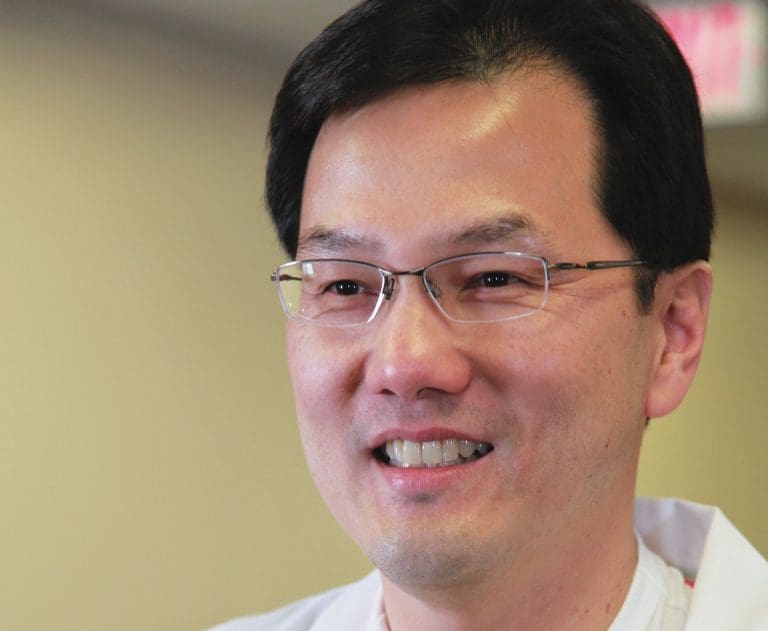 Smiling man with wire-rimmed glasses and short dark hair, wearing a white coat, in an indoor setting.