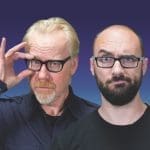 Two men posed side by side against a purple-blue backdrop, one touching his black-framed glasses, the other with a beard.