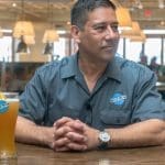 A smiling man in a gray Blue Moon shirt sits with clasped hands beside a glass of Blue Moon beer with an orange slice.