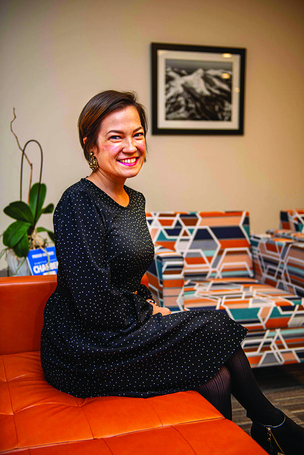 Smiling woman in a black dotted dress sits on an orange leather couch beside geometric patterned chairs.