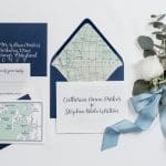 Flat-lay wedding invitation suite with navy envelopes, a map-lined liner, calligraphy cards, and white roses tied with a blue ribbon.