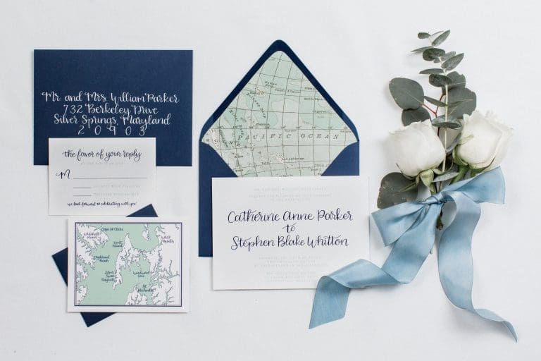 Flat-lay wedding invitation suite with navy envelopes, a map-lined liner, calligraphy cards, and white roses tied with a blue ribbon.