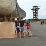 Four children stand on gravel beside a large curved wooden structure, with a tall wooden observation tower behind them.