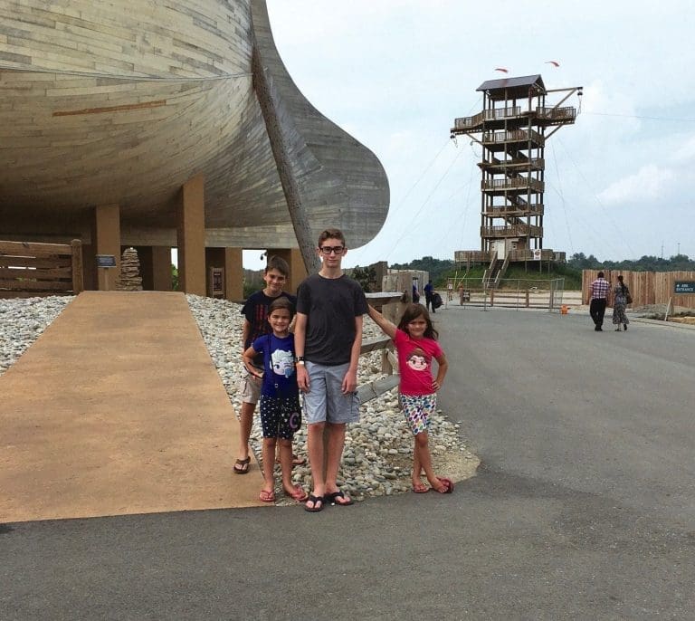 Four children stand on gravel beside a large curved wooden structure, with a tall wooden observation tower behind them.