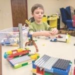 A young boy in a green shirt builds with colorful LEGO bricks at a table, with a bin of bricks nearby.