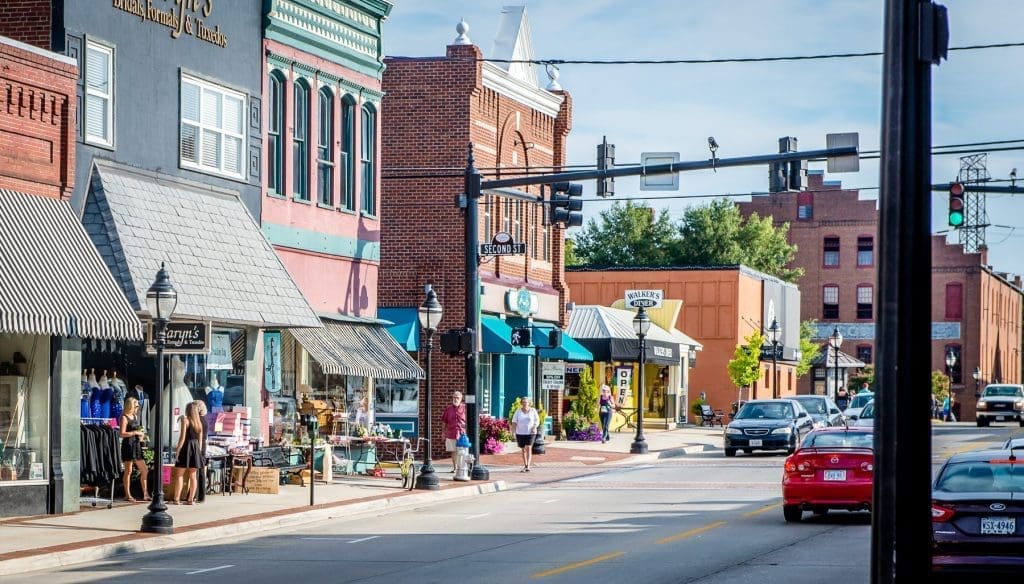 Brick storefronts with striped awnings line a sunny downtown street, with pedestrians on the sidewalk and cars driving past.