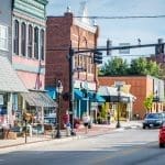 Brick storefronts with striped awnings line a sunny downtown street, with pedestrians on the sidewalk and cars driving past.