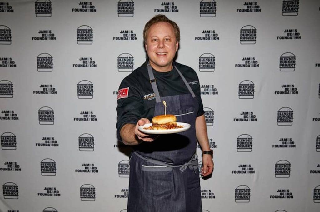 A smiling chef in a dark shirt and denim apron holds a plate with a burger in front of a James Beard Foundation Blended Burger Project backdrop.
