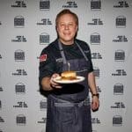 A smiling chef in a dark shirt and denim apron holds a plate with a burger in front of a James Beard Foundation Blended Burger Project backdrop.