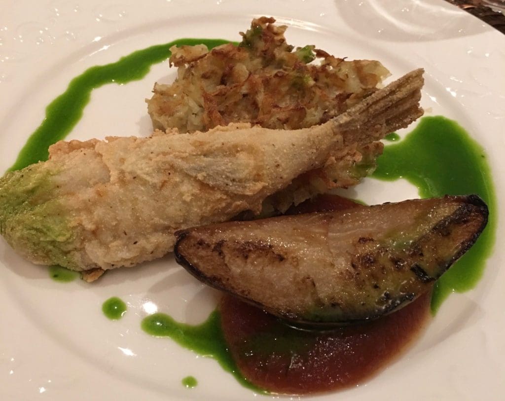 Fried whole fish with crispy shredded potato cake, a grilled mussel-like shell, and green herb sauce on a white plate.