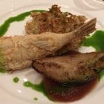Fried whole fish with crispy shredded potato cake, a grilled mussel-like shell, and green herb sauce on a white plate.