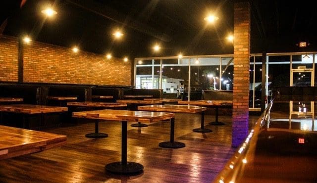 The dimly lit dining room has wooden tables, black pedestal bases, brick walls, and warm overhead string lights at night.