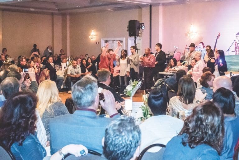 Guests in a crowded banquet hall cheer and raise their arms toward a performer on a stage, with seated diners in the foreground.