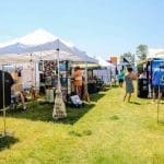 Outdoor festival vendor row under white canopy tents on grass, with shirts on racks, art displays, and shoppers strolling on a sunny day.