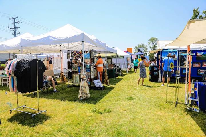 Outdoor festival vendor row under white canopy tents on grass, with shirts on racks, art displays, and shoppers strolling on a sunny day.