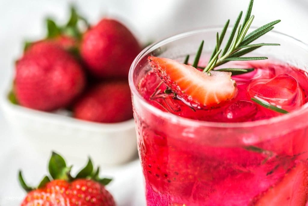 A clear glass of pink strawberry cocktail with ice, a strawberry slice, and rosemary sprig, with whole strawberries nearby.