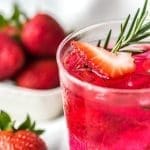 A clear glass of pink strawberry cocktail with ice, a strawberry slice, and rosemary sprig, with whole strawberries nearby.