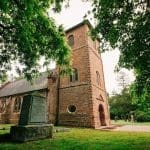 Red brick church with a tall square bell tower, arched windows, and a granite marker in a shaded churchyard.