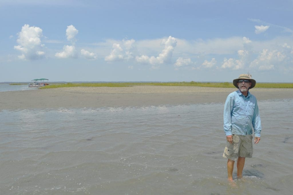 A man in a sun hat and light blue shirt stands in shallow water on a sandbar, with a pontoon boat and marsh behind him.