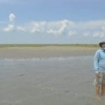 A man in a sun hat and light blue shirt stands in shallow water on a sandbar, with a pontoon boat and marsh behind him.