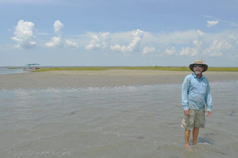 A man in a sun hat and light blue shirt stands in shallow water on a sandbar, with a pontoon boat and marsh behind him.