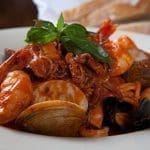 A white bowl of seafood in red tomato sauce with shrimp, clams, mussels, calamari, scallops, and fresh basil.