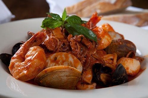 A white bowl of seafood in red tomato sauce with shrimp, clams, mussels, calamari, scallops, and fresh basil.