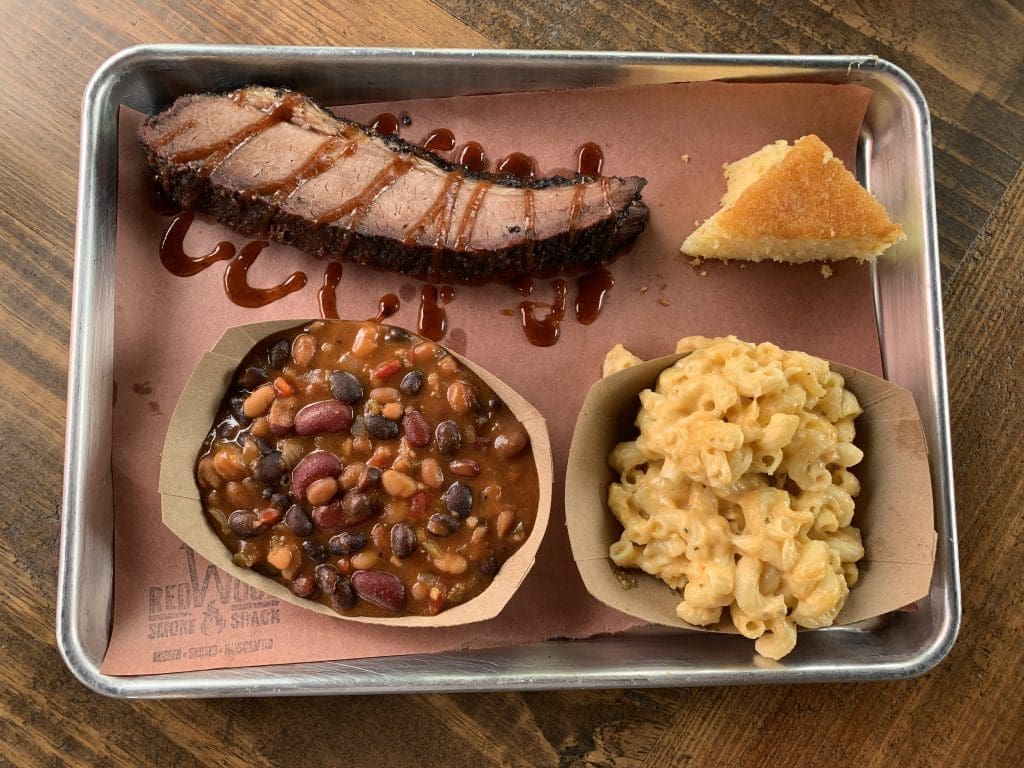 Smoked brisket slices with barbecue sauce, baked beans, macaroni and cheese, and cornbread on a metal tray, with a Red Wind Smoke & Snack logo.